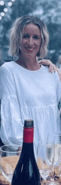 a woman in a white dress is standing next to a bottle of wine and wine glasses
