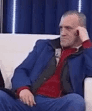 a man in a blue suit and red sweater is sitting on a couch with his hand on his chin .
