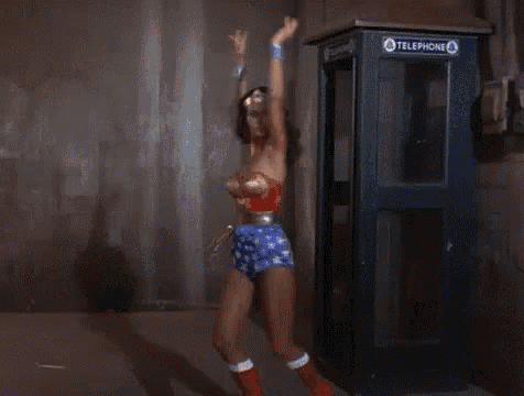 wonder woman is standing in front of a telephone booth with her arms in the air .