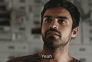 a man with a beard is standing in front of a wall and saying yeah .