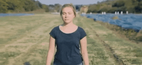 a woman in a blue t-shirt is standing in a field near a body of water .