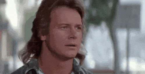 a close up of a man with long hair and a mullet looking at the camera .