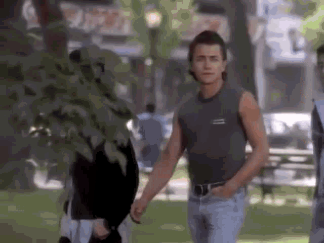 a man in a tank top and jeans is walking in a park holding a woman 's hand .