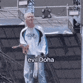 a man is standing on a roof with a microphone in his hand and the words `` evil doha '' above him .