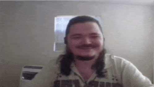 a man with long hair and a beard is smiling in front of a window