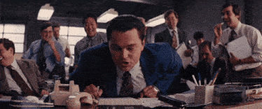 a man in a blue suit is sitting at a desk in front of a group of men in suits and ties