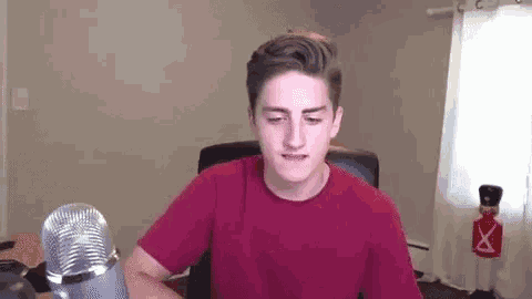 a young man in a red shirt is sitting in front of a microphone in a living room .