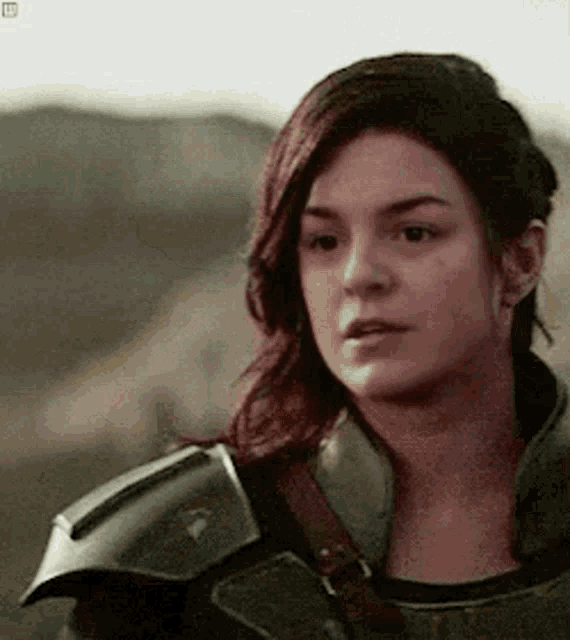 a woman with long red hair is wearing a green armored jacket and looking at the camera .
