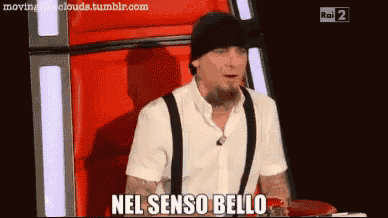a man in a white shirt and black suspenders is sitting in a chair and says " nel senso bello "