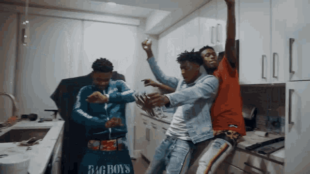 three men are dancing in a kitchen with one wearing a bag that says big boys on it