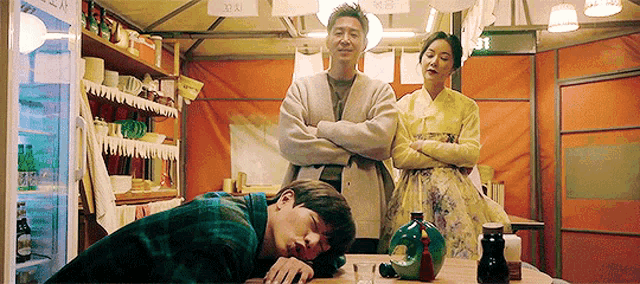 a man is sleeping on a table while a woman and a man stand behind him .