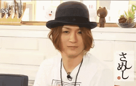 a man wearing a bowler hat and a white shirt is sitting in front of a shelf that says 5:55