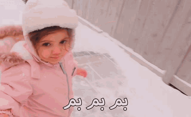a little girl wearing a pink jacket and a white hat is waving her hand in the snow