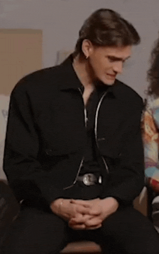 a man in a black jacket is sitting down with his hands folded .