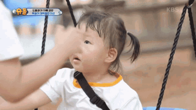 a little girl is sitting on a swing with a kbs logo in the corner