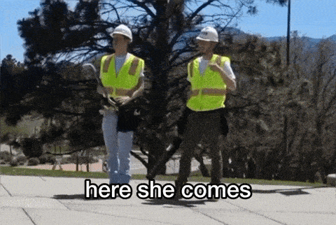 two construction workers wearing hard hats and safety vests are standing on a sidewalk and one of them says here she comes .