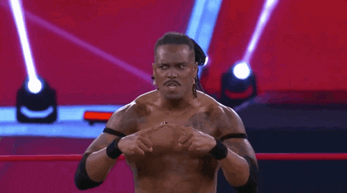 a wrestler is standing in a ring with his hands on his chest and a referee behind him .