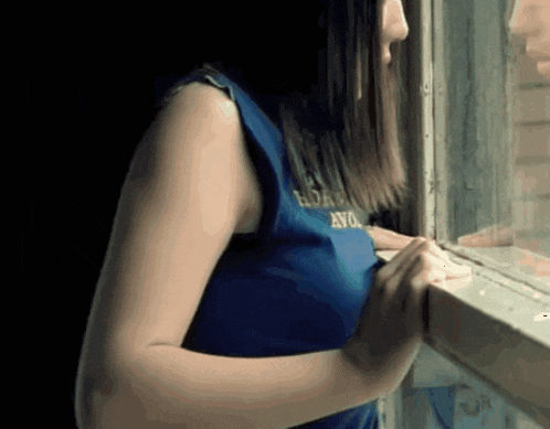 a woman in a blue shirt with the word ayo on it is looking out of a window