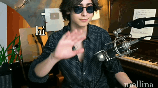 a young man wearing sunglasses and a black shirt is sitting in front of a microphone saying hi