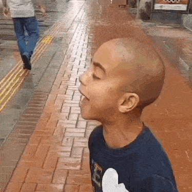 a young boy with a bald head is standing on a brick sidewalk with his mouth open .