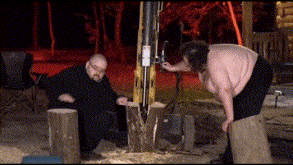 a man is standing next to a woman who is cutting a tree stump with a saw .