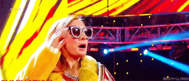 a woman wearing sunglasses and a fur coat is standing on a stage in front of a colorful background .