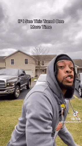 a man in a hoodie is standing in front of a house with cars parked in front of him .