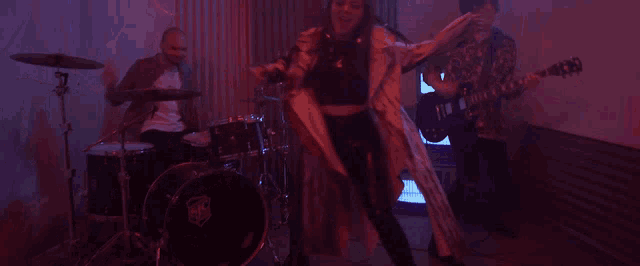 a woman is dancing in front of a drum set and a man playing guitar