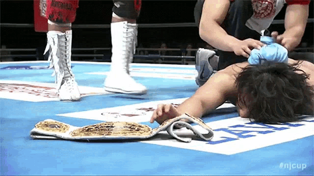 a wrestler is laying on the ground in a wrestling ring while another wrestler stands behind him .
