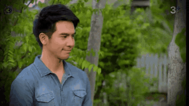a man in a blue denim shirt is standing in front of a tree .