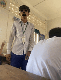 a young man with a mask on his face stands in a classroom