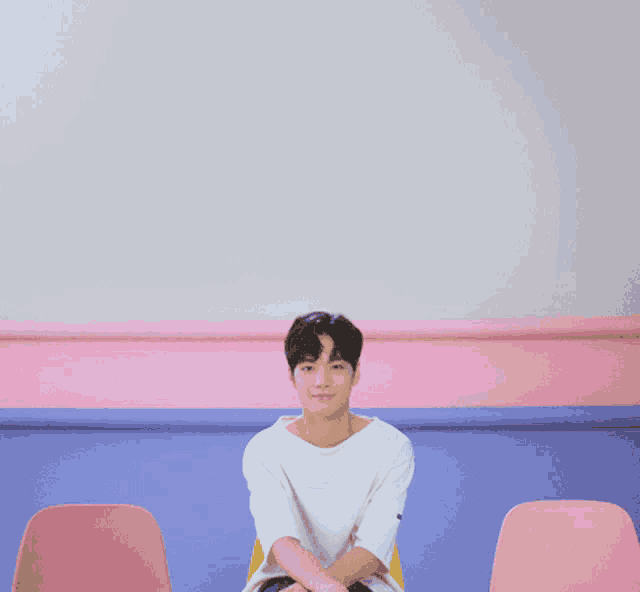 a young man in a white shirt sits in front of a pink and blue wall
