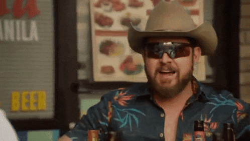 a man wearing a cowboy hat and sunglasses is sitting at a table holding a bottle of beer .