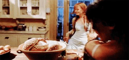 a woman in a white dress is standing in a kitchen with a bowl of bread on the table .