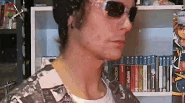 a man wearing a hat and sunglasses is standing in front of a bookshelf .