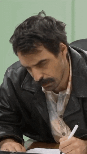 a man with a mustache is sitting at a desk writing on a piece of paper