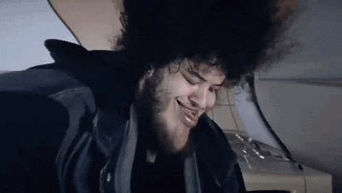 a man with curly hair and a beard is sitting on a plane with his head in the air .