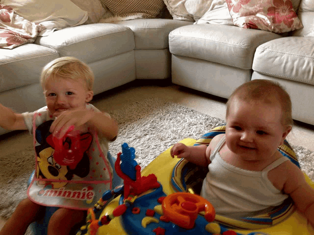 a baby wearing a minnie apron sits next to a baby in a walker