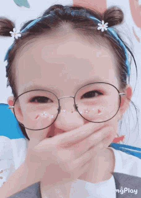 a little girl wearing glasses and a flower in her hair covering her mouth