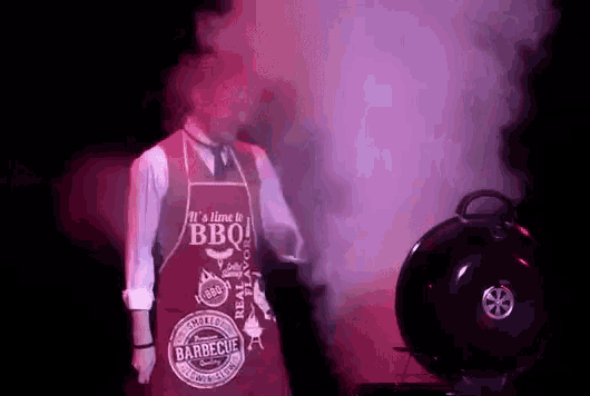 a man wearing a red apron is standing in front of a barbecue grill .