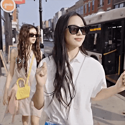 two women wearing sunglasses are walking down a sidewalk next to a bus