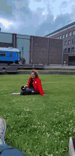 a woman in a red shirt sits on the grass in front of a blue train