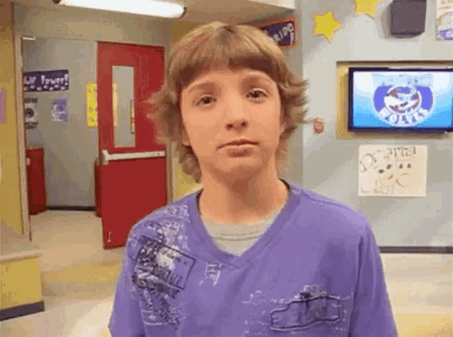 a young boy in a purple shirt is standing in a hallway with a tv on the wall .