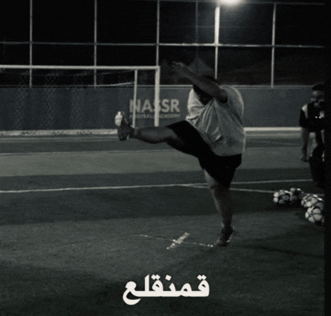 a man kicks a soccer ball on a field with nasr written on the fence behind him