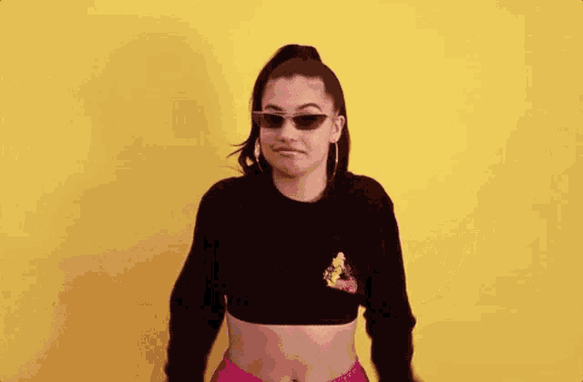 a woman wearing sunglasses and a crop top is standing in front of a yellow wall .