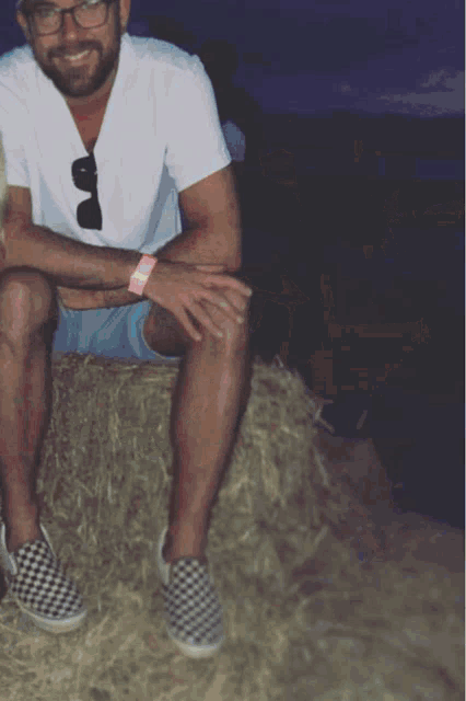 a man in a white shirt and blue shorts sits on a bale of hay with his legs crossed