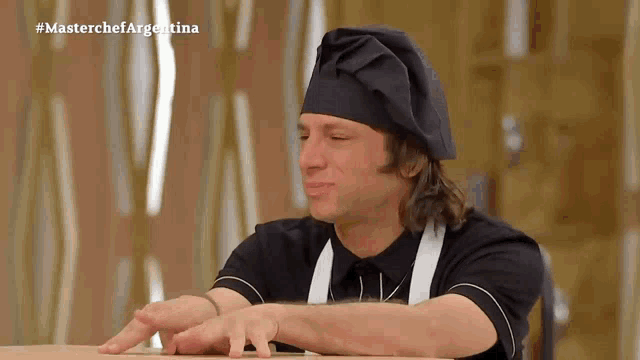 a man wearing a chef 's hat and apron is sitting at a table with #masterchefargentina written on the screen