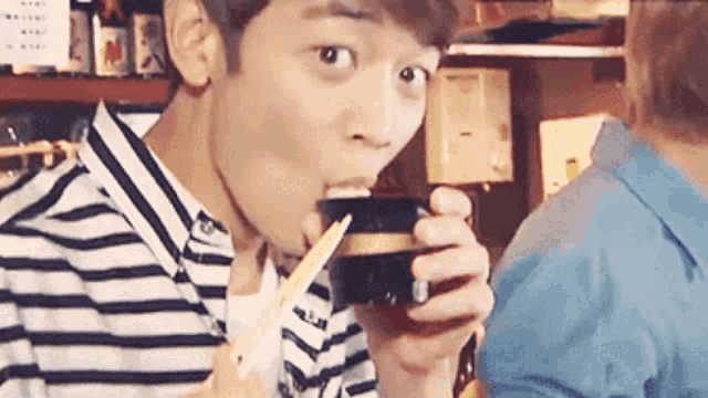 a young man in a striped shirt is drinking from a bowl with chopsticks