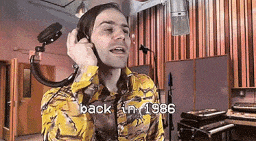 a man in a yellow shirt is wearing headphones and singing into a microphone in a studio .