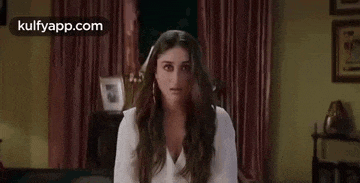 a woman in a white dress is sitting in a living room and looking at the camera .